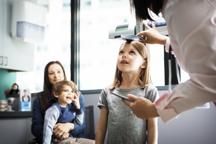 Une mère est assise sur un fauteuil avec l'un de ses enfants sur les genoux. Sa petite fille est en train de se faire osculté devant elle, chez un cabinet de pédiatre.